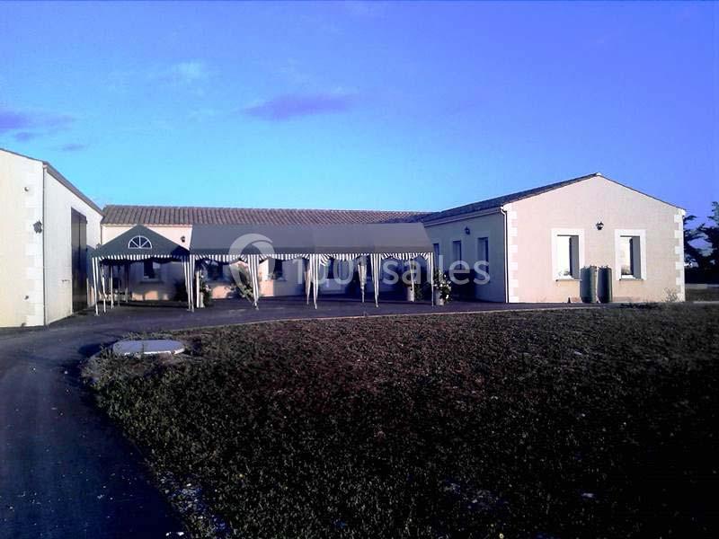 Bâtiment clair de plain-pied avec une tonnelle rayée à l'entrée, entouré de pelouse et d'un chemin asphalté.