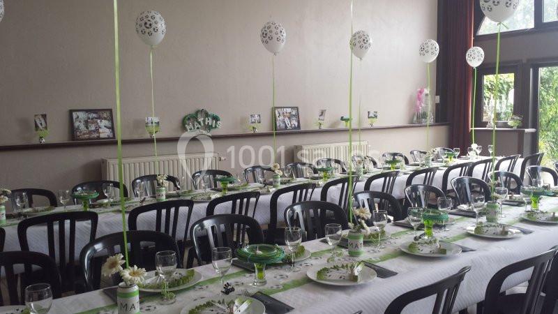 Salle décorée pour une réception avec longues tables dressées, ballons blancs suspendus et décoration verte.