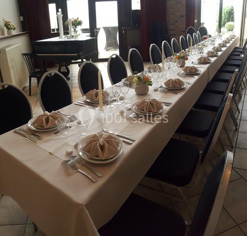 Table dressée avec nappes blanches, assiettes, verres, serviettes pliées et bougies, dans une salle lumineuse.