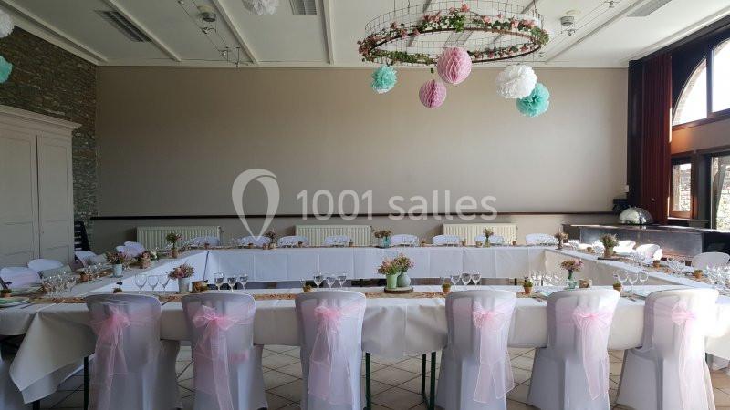 Salle décorée pour un événement, avec des tables en U, nappes blanches, chaises ornées de rubans roses et décorations…
