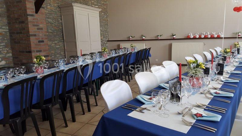 Salle décorée pour un repas, avec longues tables dressées, nappes bleues, chaises alternées noires et blanches, et décor…