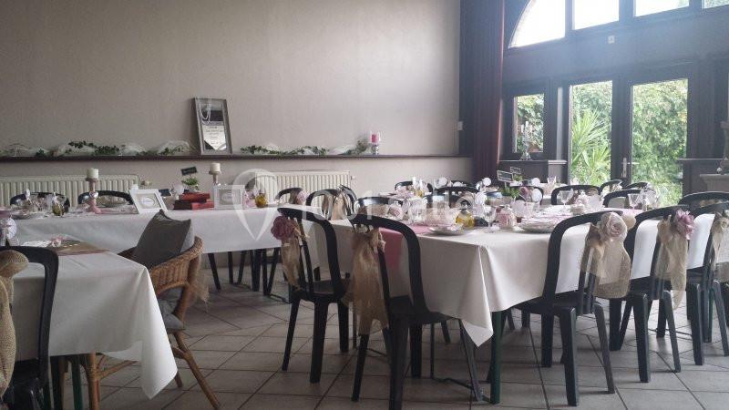 Salle décorée pour un événement avec des tables dressées, nappes blanches, chaises ornées et lumière naturelle.