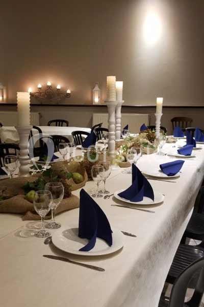 Table dressée avec nappes blanches, serviettes bleues pliées, bougies et décoration végétale dans une salle éclairée.