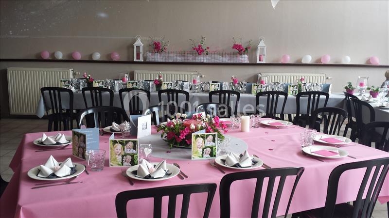 Salle décorée pour un événement, avec tables dressées, nappes roses, centres de table fleuris et chaises noires.