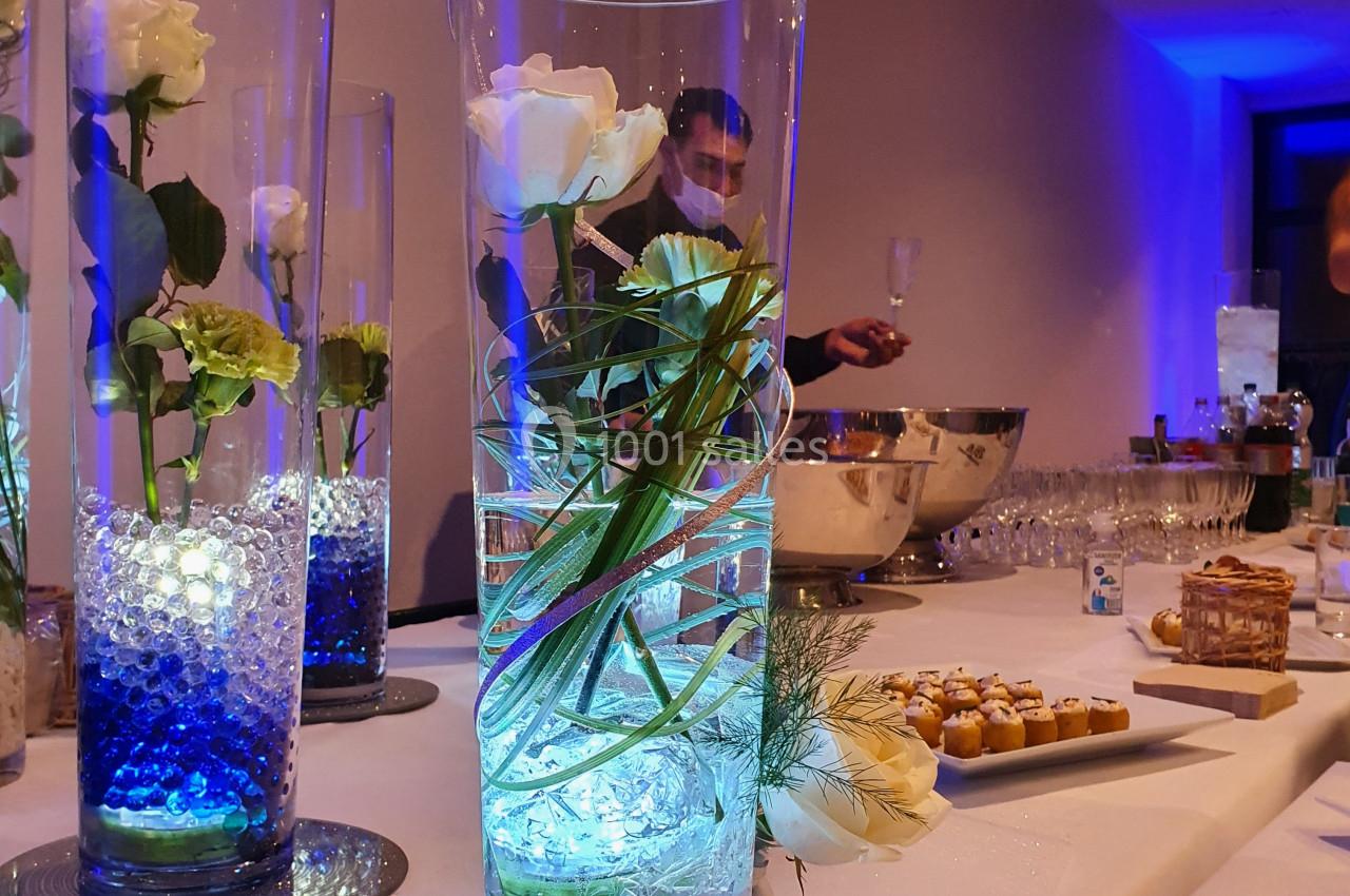 Vase en verre contenant une rose blanche et des perles lumineuses bleues, posé sur une table de réception.