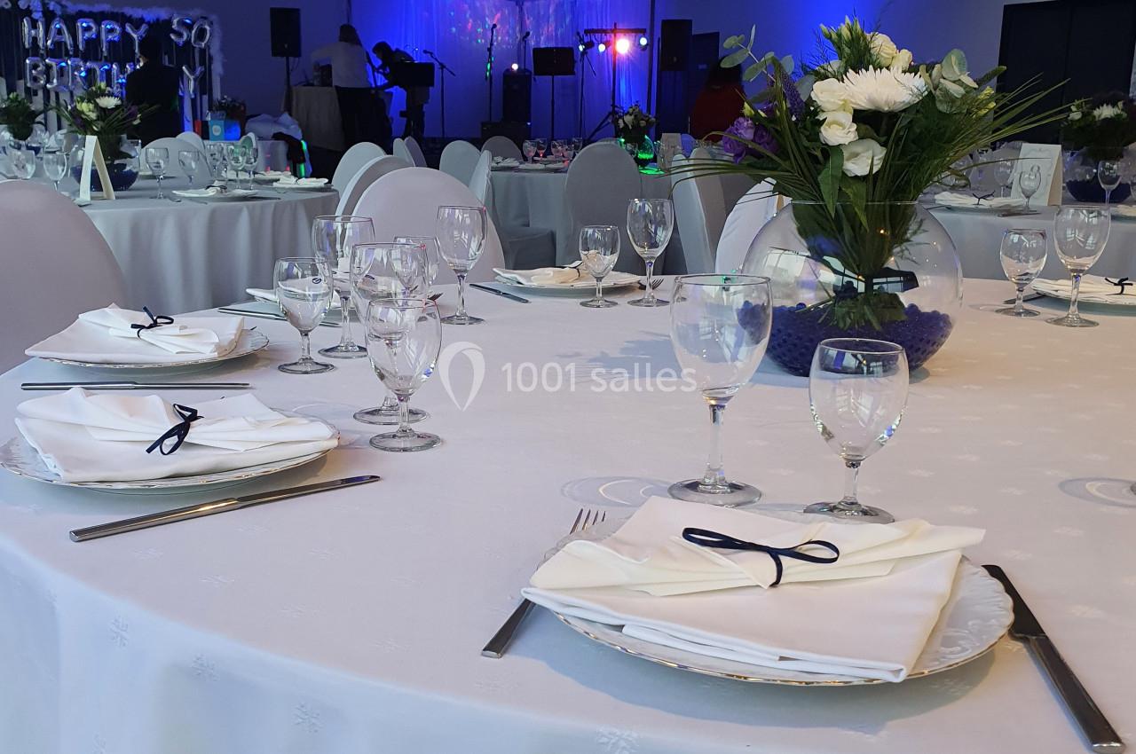 Salle de réception décorée avec des tables dressées, nappes blanches, fleurs et éclairage d'ambiance bleu.