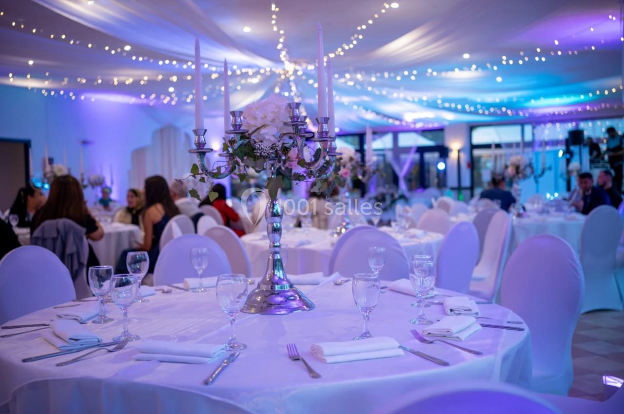 Salle de réception décorée avec des tables rondes, nappes blanches, chandeliers et guirlandes lumineuses au plafond.