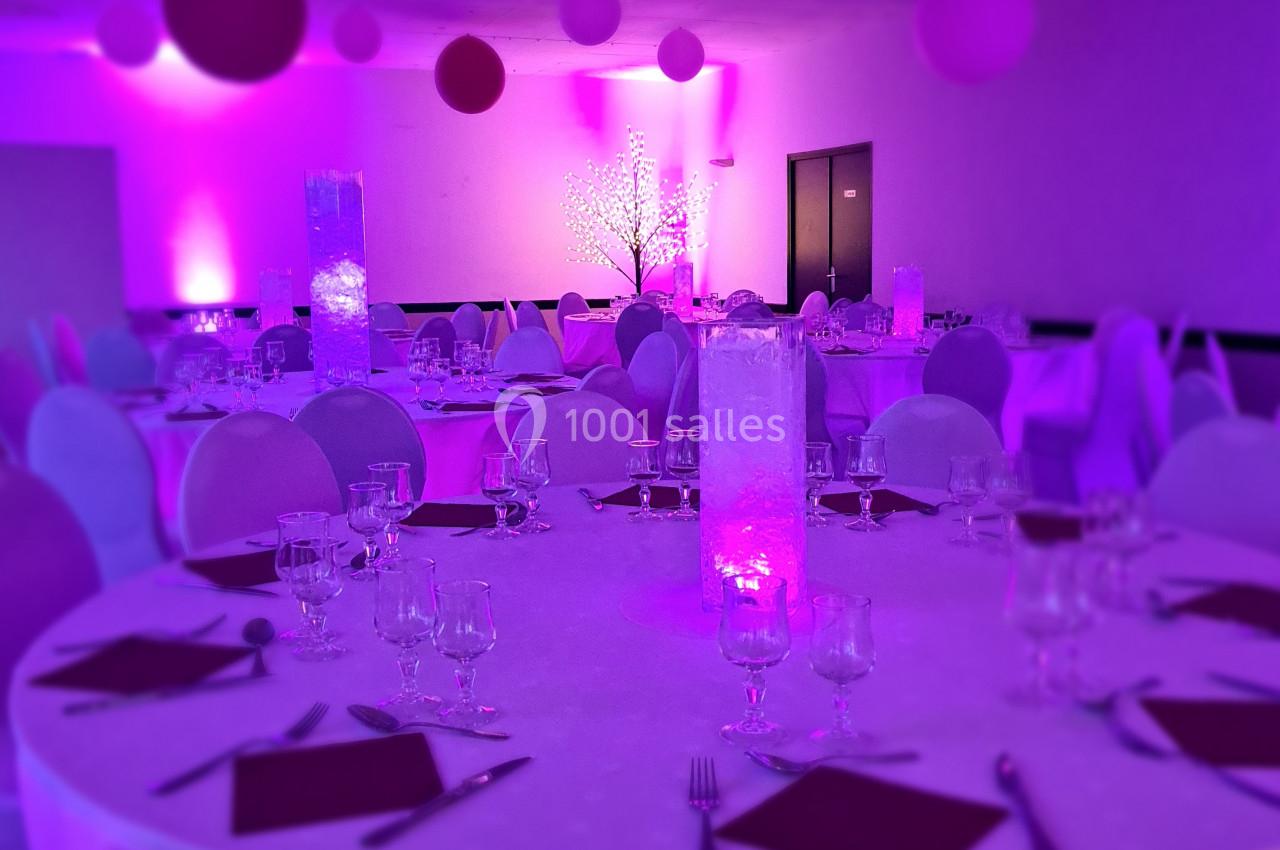 Salle décorée pour un événement avec tables rondes, nappes blanches, éclairage rose et ballons suspendus.