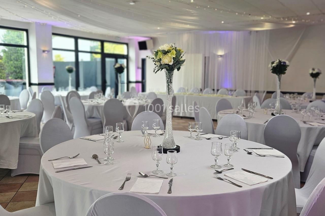 Salle de réception élégante avec tables rondes dressées, nappes blanches et décorations florales au centre.