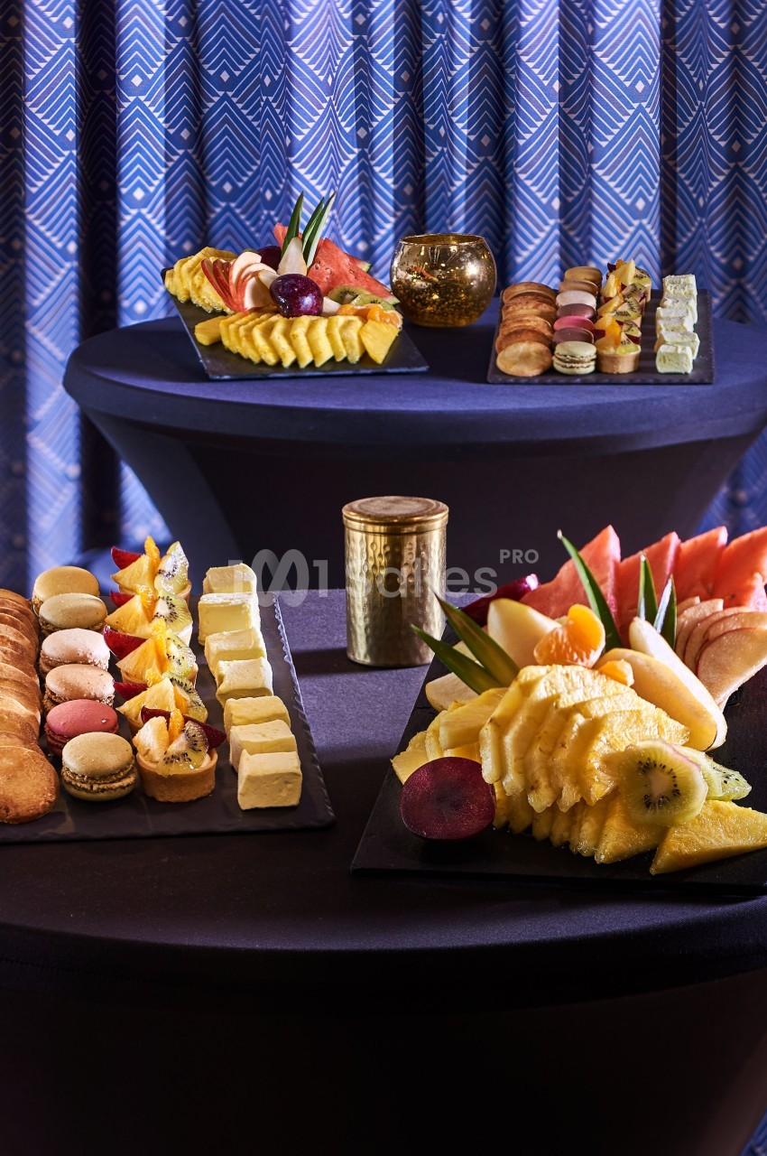 Plateaux de fruits frais, pâtisseries et macarons disposés sur des tables drapées de noir devant un rideau à motifs.