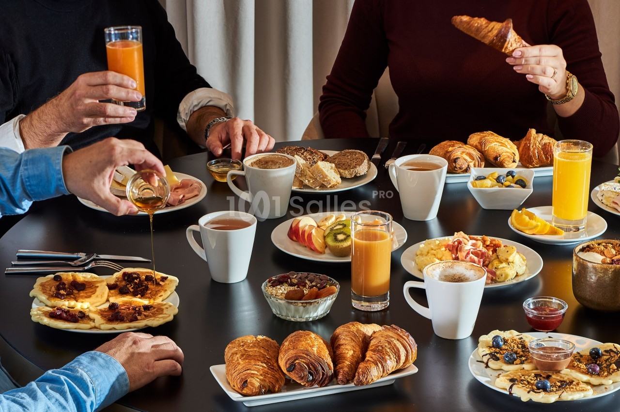 Petit-déjeuner varié avec viennoiseries, fruits, pancakes, boissons chaudes et jus d'orange sur une table noire.