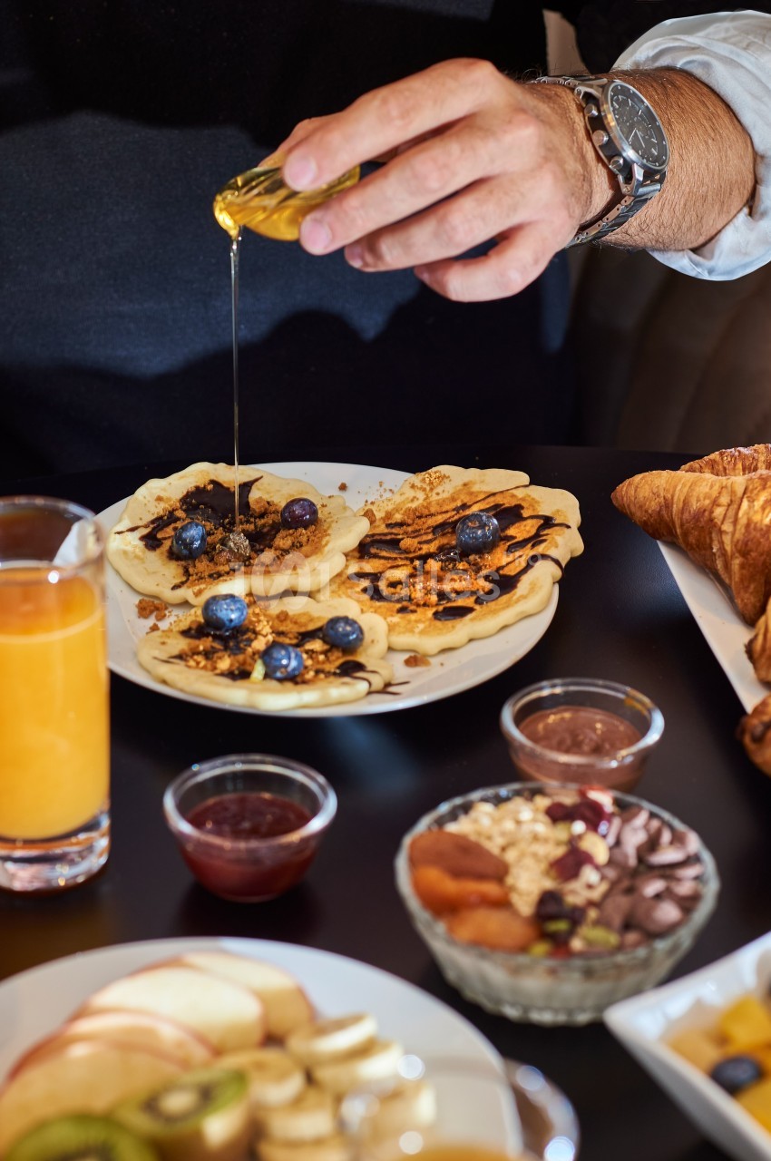 Une main verse du miel sur des pancakes garnis de myrtilles et de noix, entourés de divers aliments pour le petit-déjeuner.