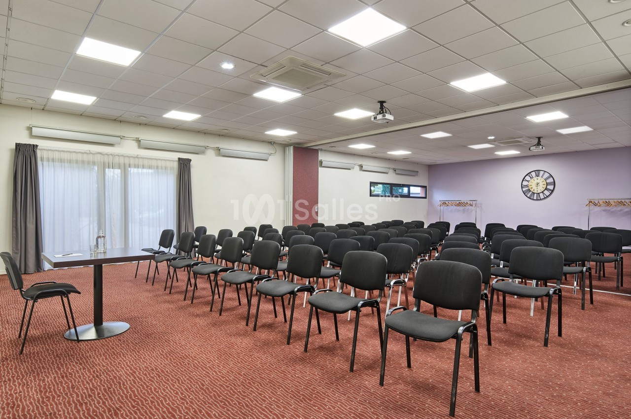 Salle de conférence avec des rangées de chaises noires, une table à gauche et un mur avec une horloge en fond.