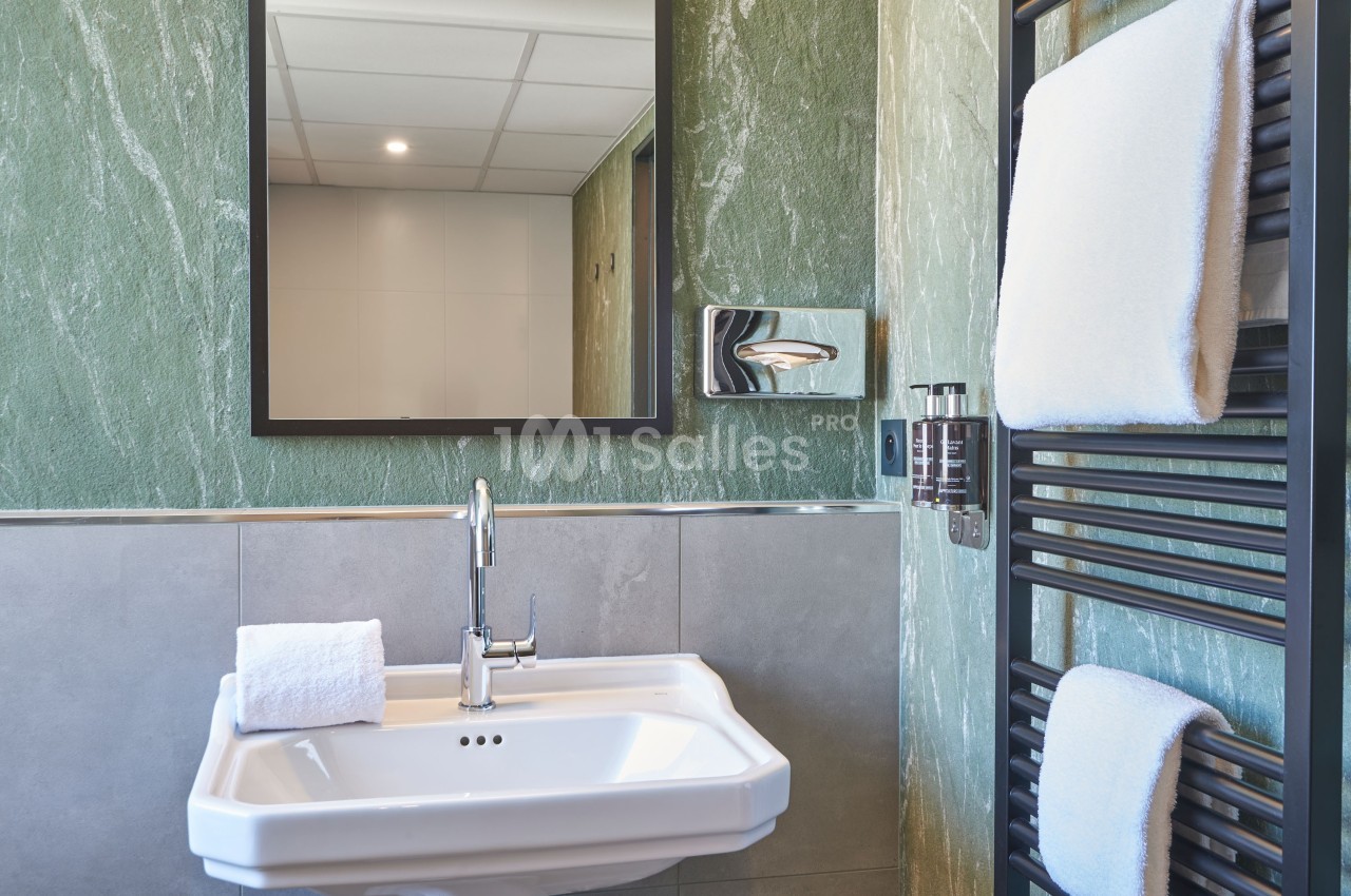 Lavabo blanc avec miroir rectangulaire, mur vert texturé, porte-serviettes noir et serviettes blanches.