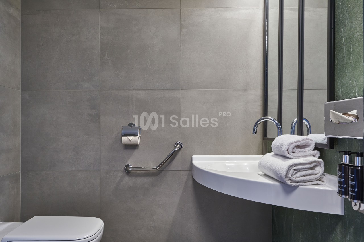 Salle de bain moderne avec lavabo, serviettes pliées, barre d'appui près des toilettes et murs en carrelage gris.