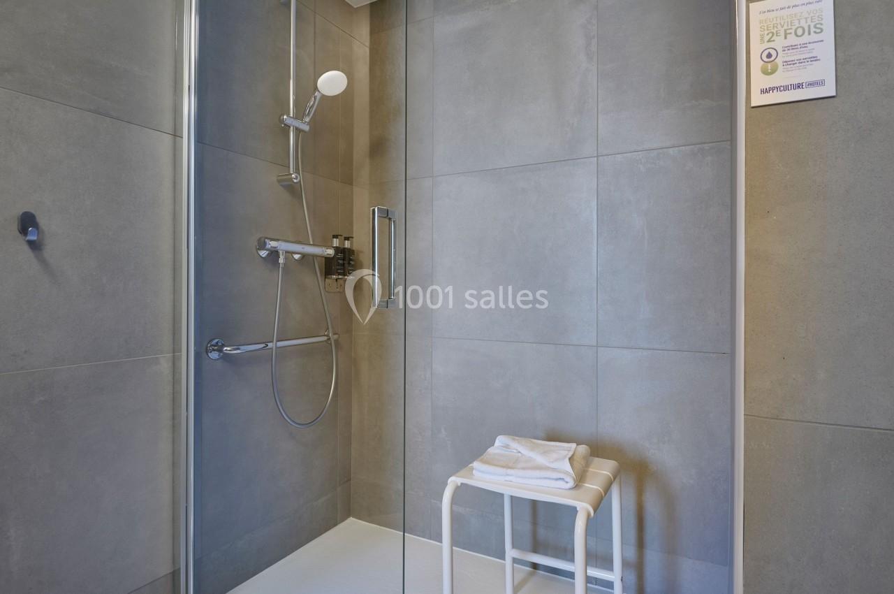 Douche moderne avec parois en verre, carrelage gris, pommeau mural et tabouret blanc posé au sol.