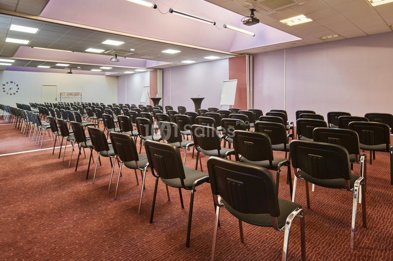 Salle de conférence vide avec des rangées de chaises noires alignées sur un sol rouge, éclairée par des plafonniers.