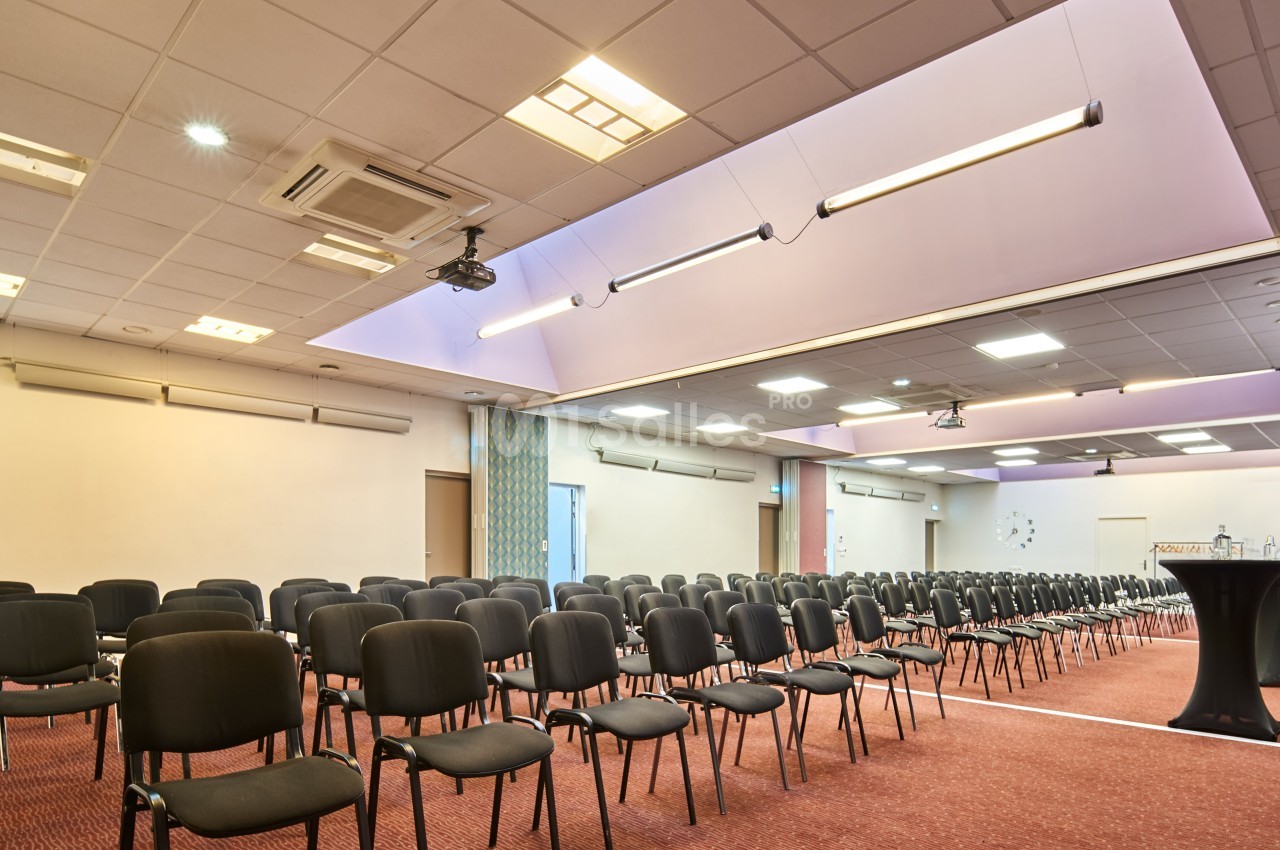 Salle de conférence vide avec rangées de chaises noires, éclairage au plafond et projecteur suspendu.