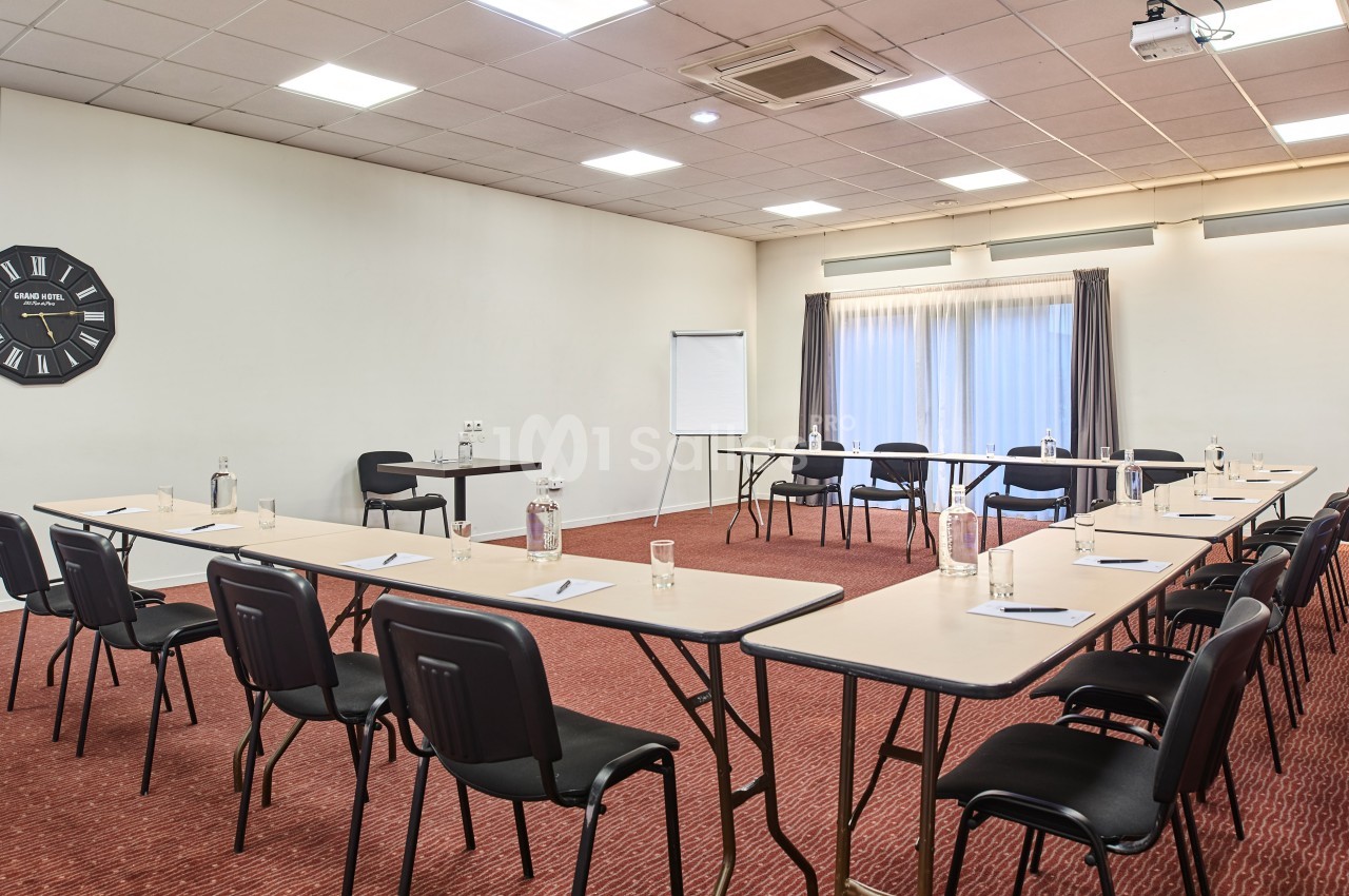 Salle de réunion lumineuse avec disposition en U, tables, chaises, tableau blanc et bouteilles d'eau sur tapis rouge.