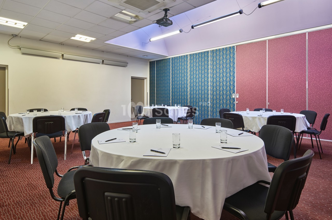 Salle de réunion avec tables rondes dressées de nappes blanches, chaises noires et fournitures de bureau.