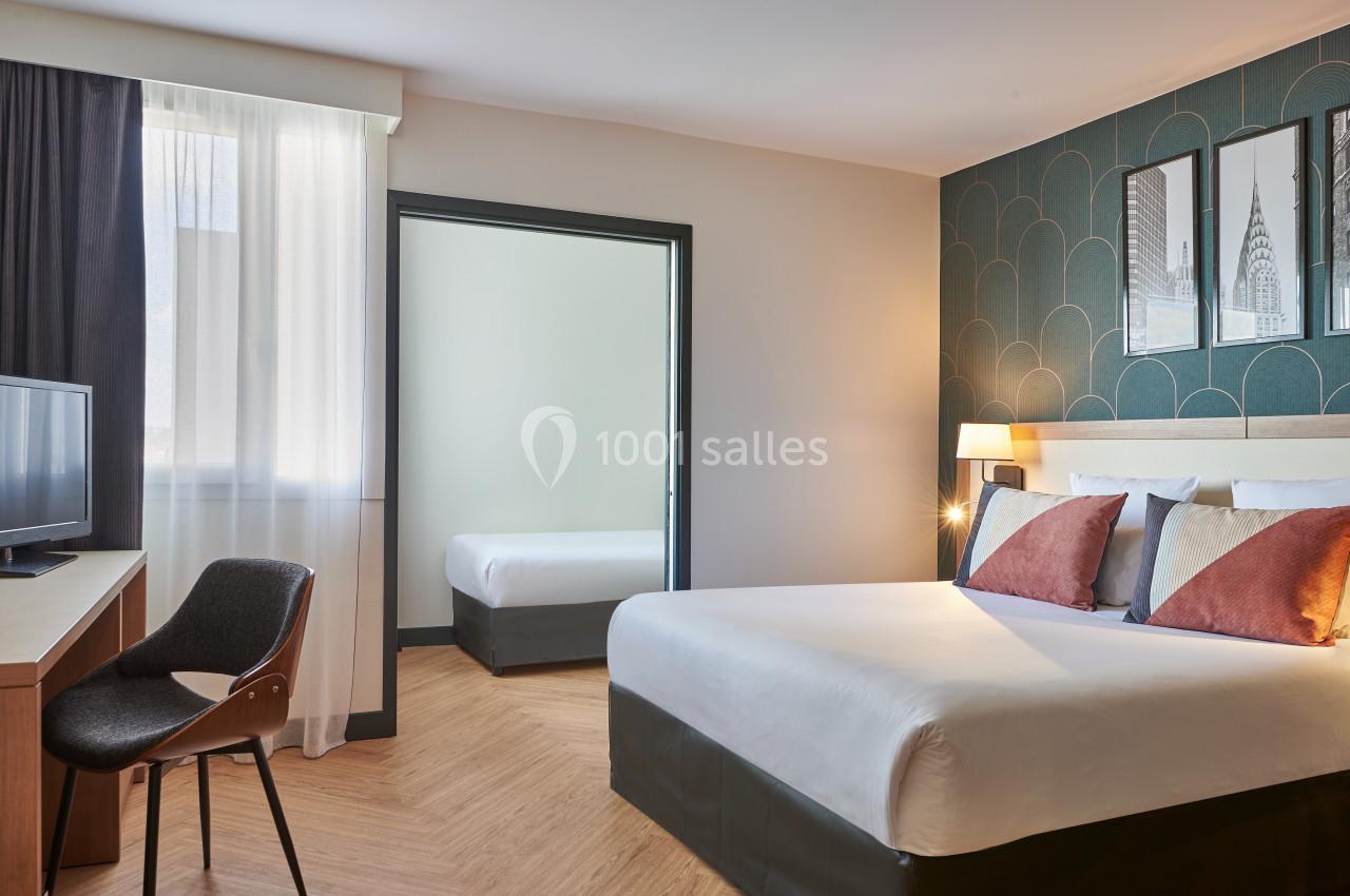 Chambre d'hôtel lumineuse avec un grand lit, un bureau, une chaise, et une pièce attenante visible à travers une porte…