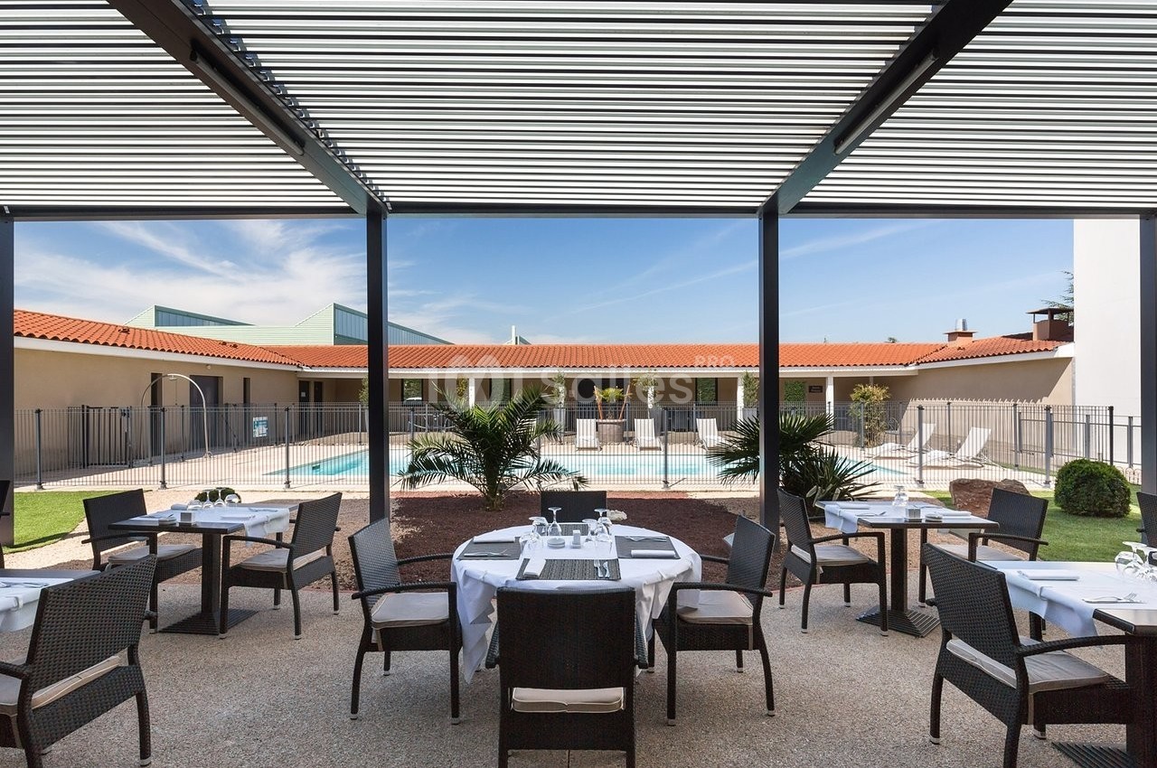 Terrasse aménagée avec des tables et des chaises, donnant sur une piscine entourée de végétation et de bâtiments.