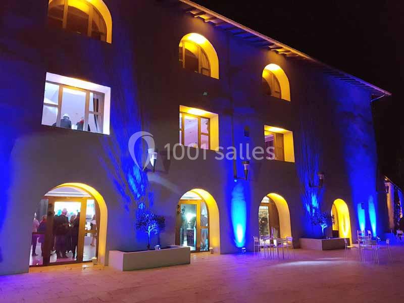 Façade d'un bâtiment éclairé en bleu et jaune la nuit, avec des fenêtres illuminées et des tables en extérieur.