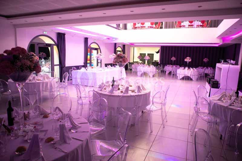 Salle de réception décorée avec des tables rondes, nappes blanches, chaises transparentes et éclairage rose.
