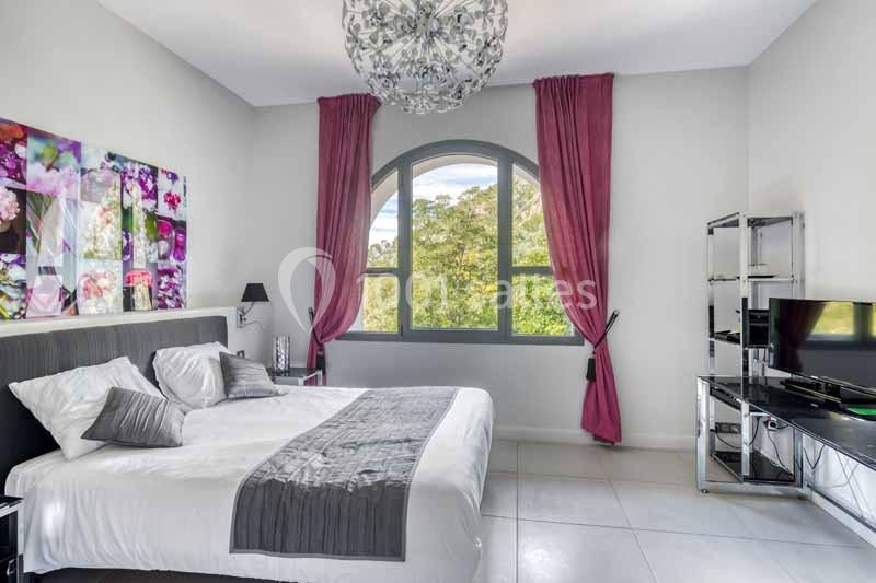 Chambre lumineuse avec lit double, rideaux roses, tableau floral, grande fenêtre cintrée et mobilier moderne.