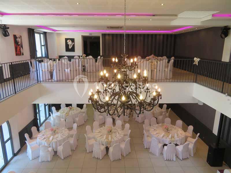 Salle de réception lumineuse avec tables rondes dressées et grand lustre central, vue depuis une mezzanine.