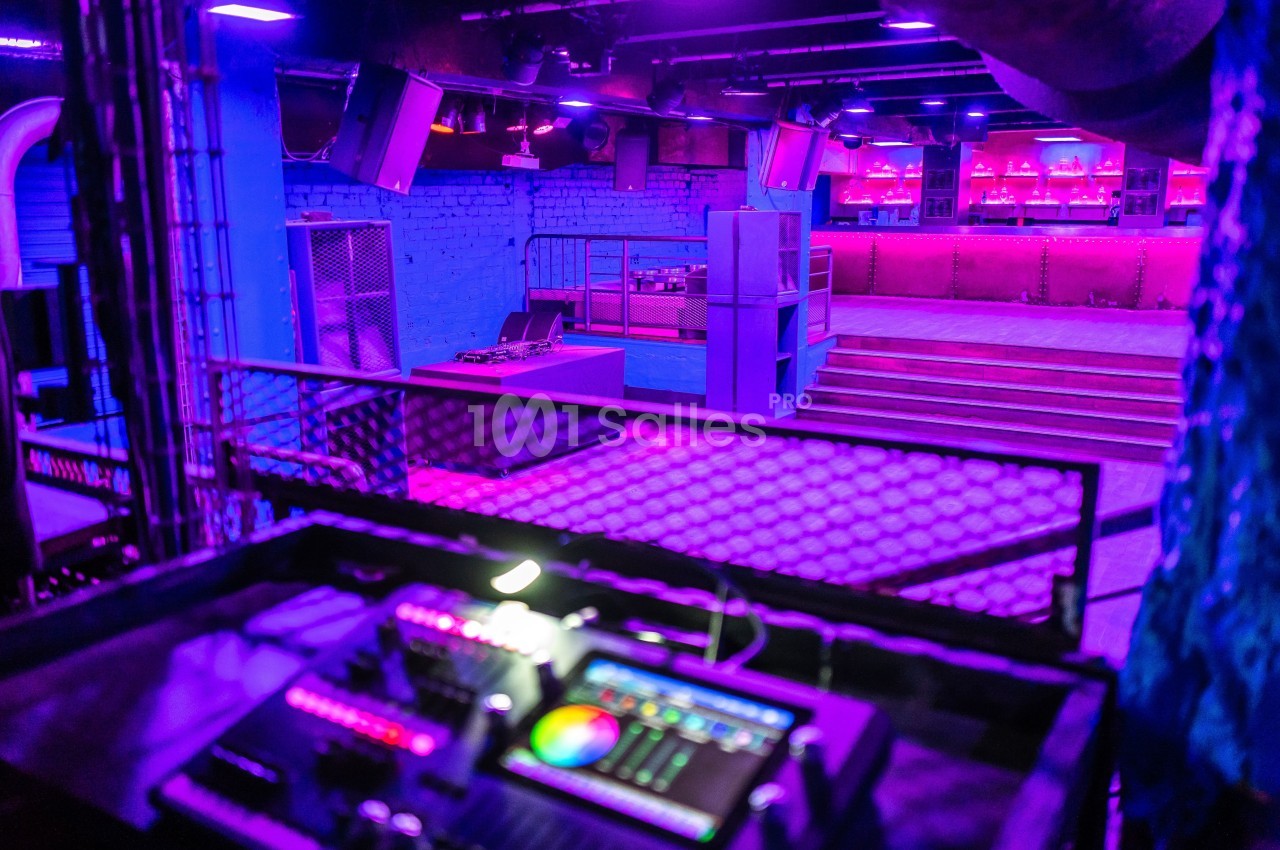 Salle de concert vide avec éclairage violet, une console de mixage au premier plan et un bar en arrière-plan.