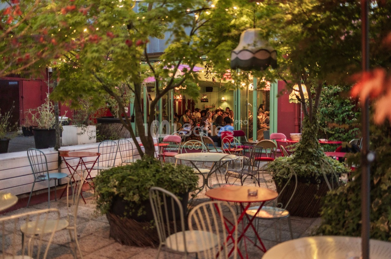 Terrasse extérieure avec tables et chaises entourées de plantes, éclairée par des guirlandes lumineuses en soirée.