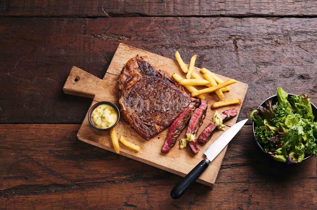 Steak grillé avec frites, sauce et salade, présenté sur une planche en bois avec un couteau sur une table rustique.