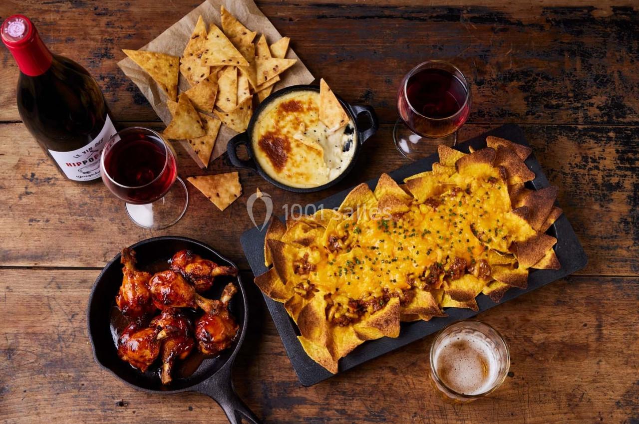 Assortiment de nachos, ailes de poulet, fromage fondu, chips et boissons sur une table en bois.