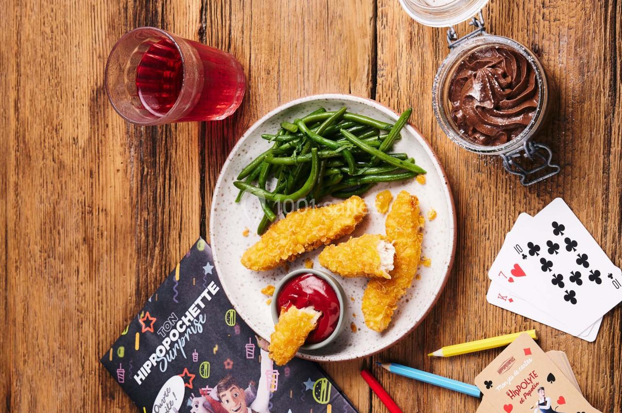 Assiette avec bâtonnets de poulet panés, haricots verts, sauce ketchup, verre de boisson rouge et mousse au chocolat.