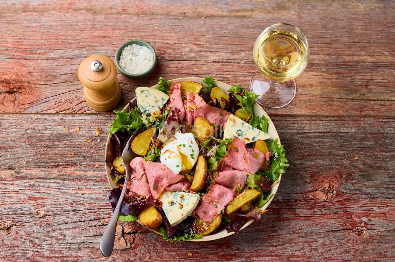 Assiette de salade composée avec rosbif, pommes de terre, fromage bleu, œuf poché et herbes, sur table en bois.