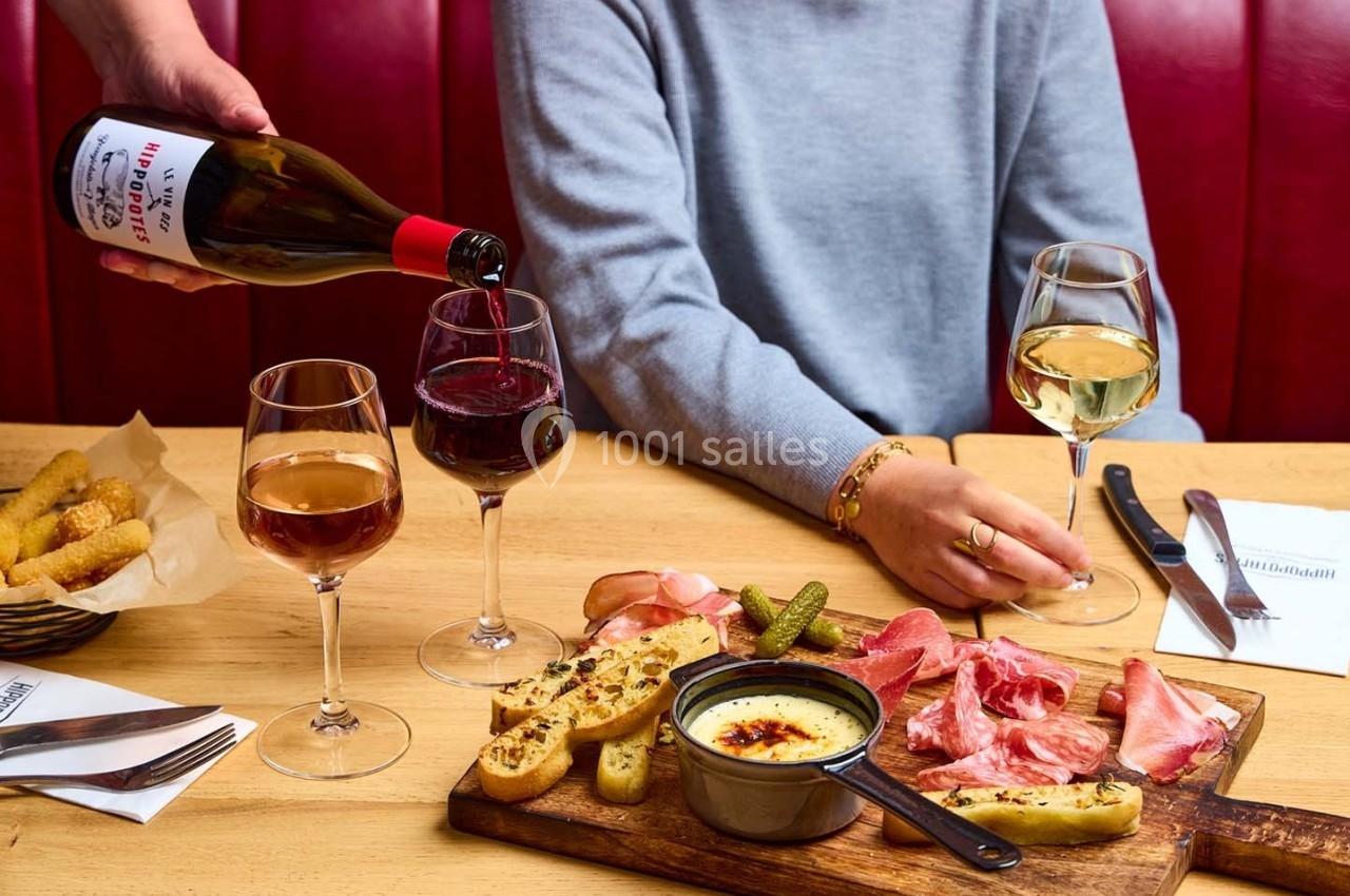 Un serveur verse du vin rouge dans un verre sur une table garnie de charcuterie, fromage fondu et autres accompagnements.