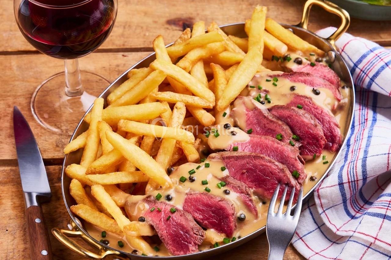 Plat de viande rouge tranchée avec sauce au poivre, accompagné de frites, servi dans un plat ovale sur une table en bois.