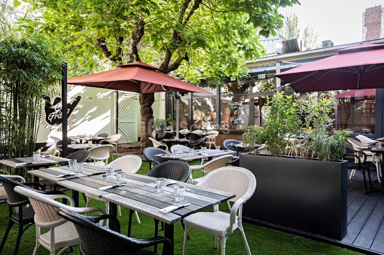 Terrasse extérieure avec tables en bois, chaises variées, parasols rouges et verdure environnante.