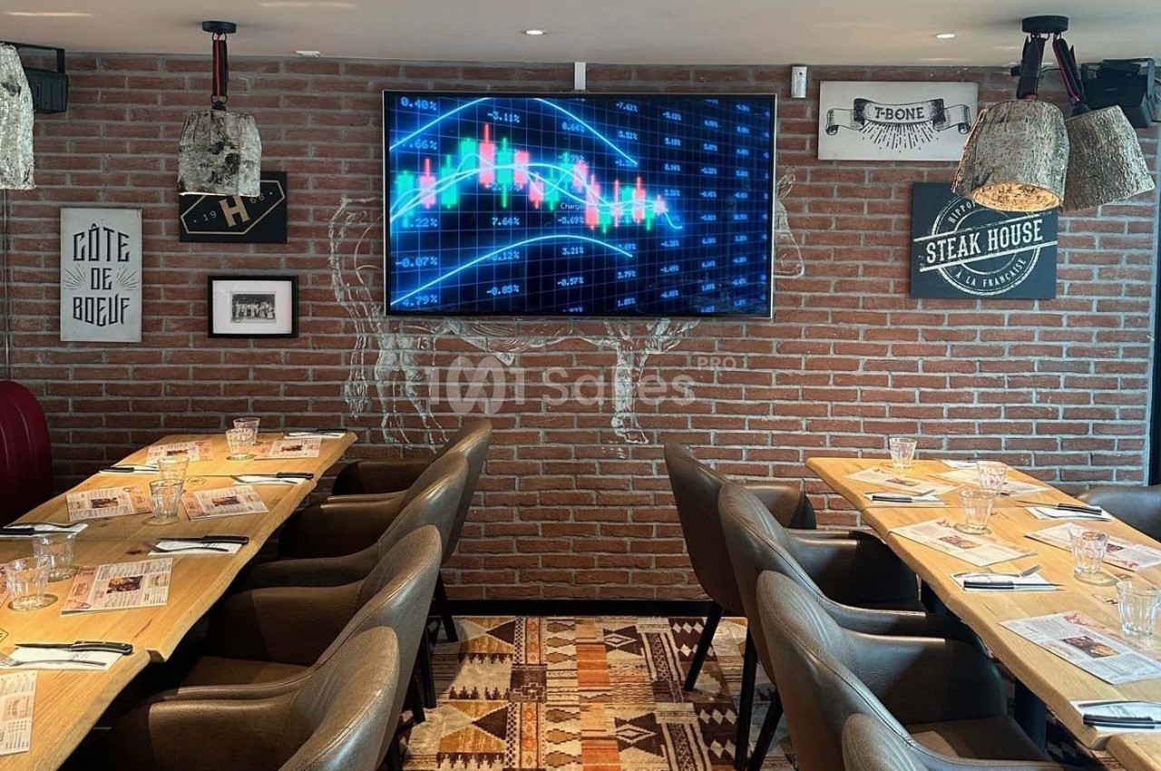 Salle de restaurant avec tables dressées, écran affichant un graphique financier et murs en briques décorés.