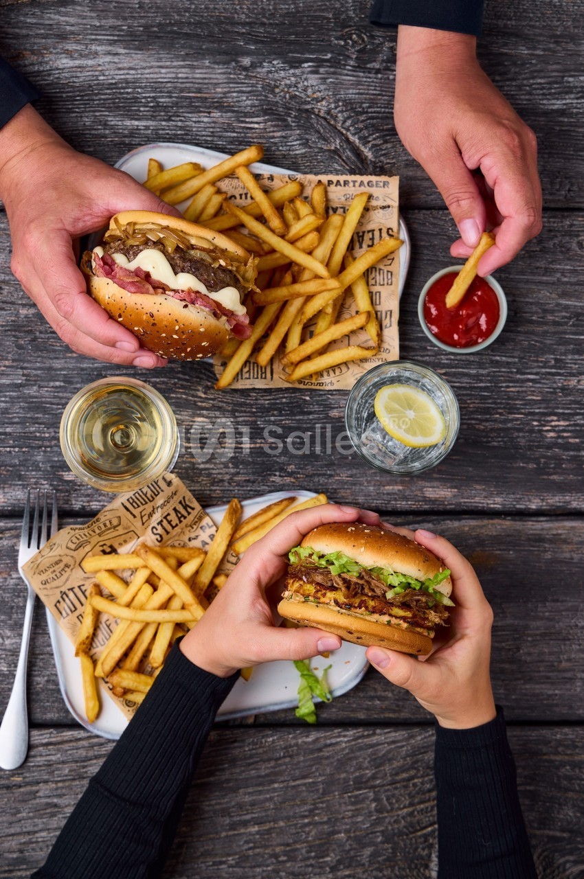 Deux personnes dégustent des burgers accompagnés de frites et de boissons sur une table en bois.