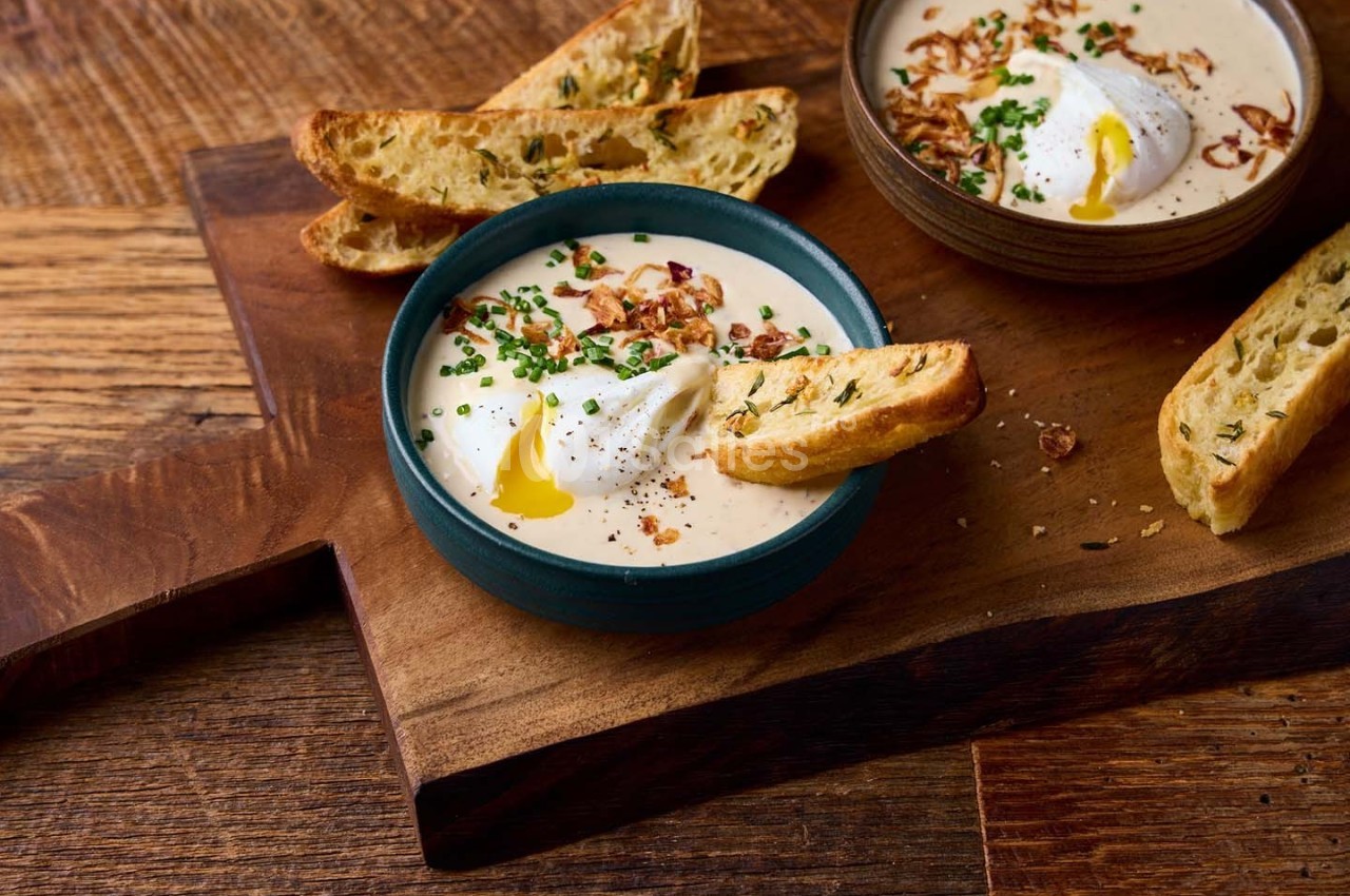Bol de soupe crémeuse garnie de ciboulette, œufs pochés et croûtons, posé sur une planche en bois.