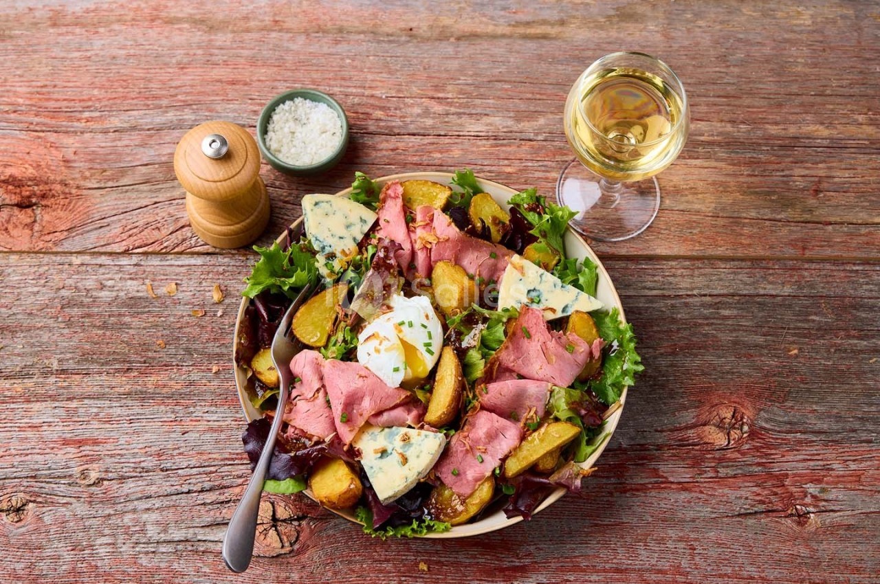 Assiette de salade composée avec rosbif, pommes de terre rôties, fromage bleu et herbes, accompagnée de vin blanc.