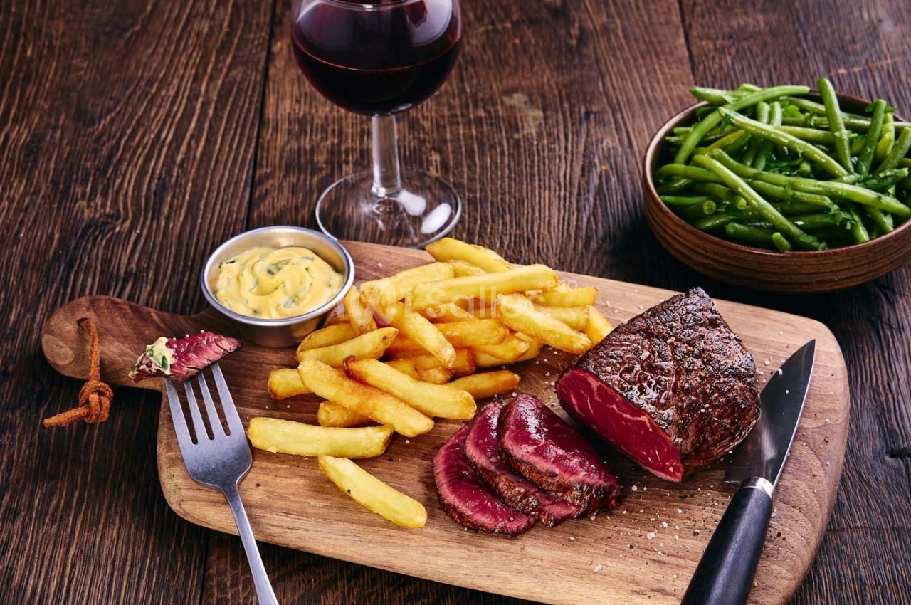 Assiette en bois avec un steak saignant tranché, des frites, une sauce, un verre de vin rouge et des haricots verts.