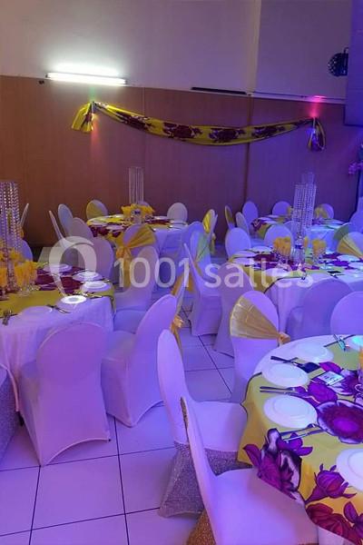 Salle de réception décorée avec des tables rondes, nappes jaunes à motifs floraux violets et chaises blanches.