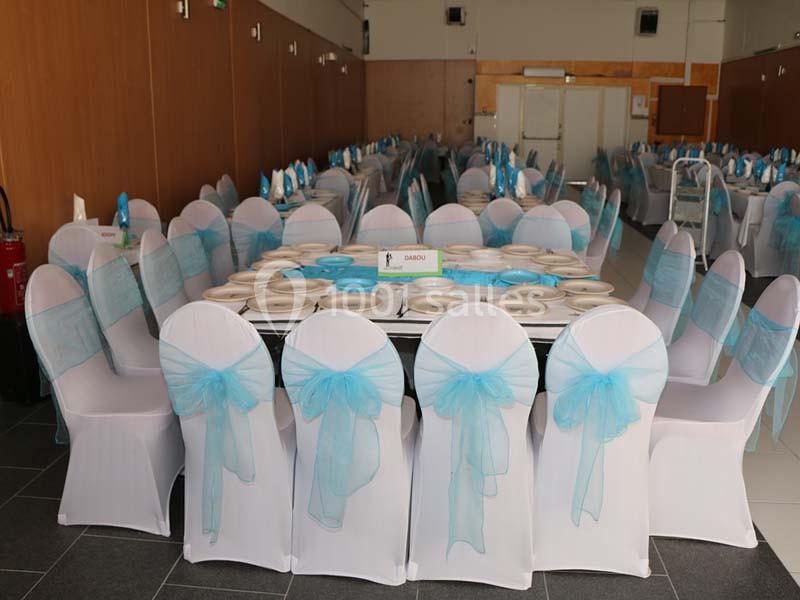 Salle de réception avec des tables dressées, chaises blanches décorées de nœuds bleus et vaisselle disposée.