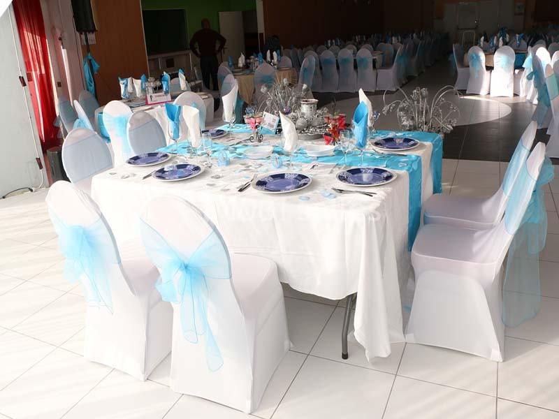 Table dressée avec nappes blanches, assiettes bleues et chaises couvertes de housses blanches ornées de nœuds bleus.