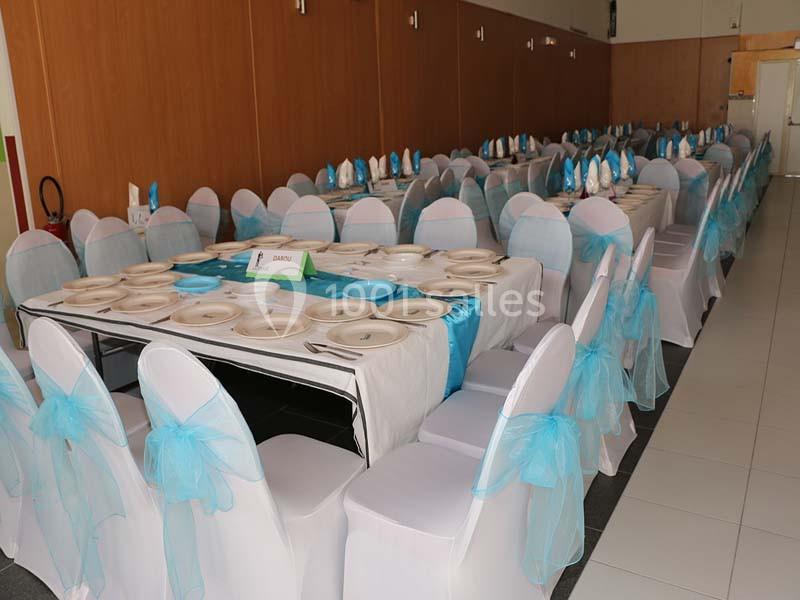 Salle de réception avec tables dressées, nappes blanches, serviettes bleues et chaises décorées de nœuds bleus.