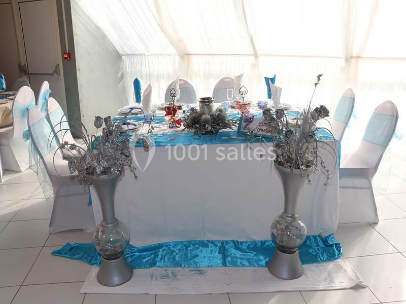 Table décorée avec nappes blanches et bleues, ornée de centres de table argentés, entourée de chaises habillées.