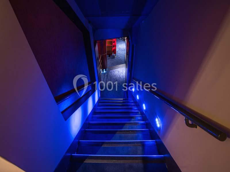 Escalier étroit éclairé par des lumières bleues, menant à une salle avec des sièges rouges visibles au fond.